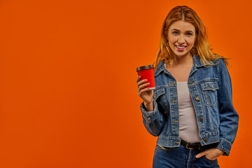 A woman with wavy fair hair in a denim jacket smiles with a red cardboard cup of coffee in her hand.