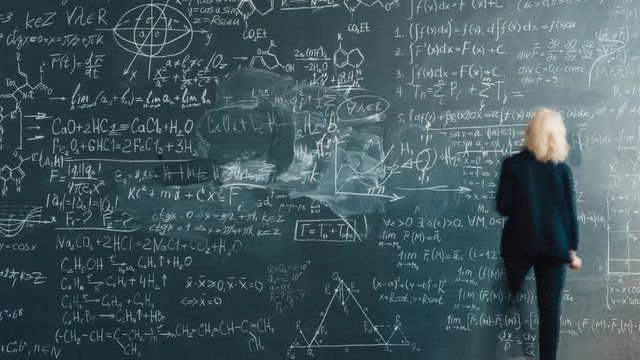 Zoom-in time lapse of intelligent female teacher in suit writing formlas on blackboard with chalk teaching science. People and modern education concept.