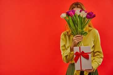 Happy girl in a yellow blouse play with a bouquet of tulips and showes a white box tied with red ribbon.