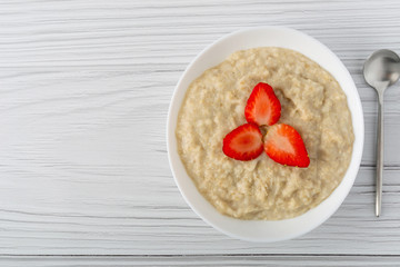 Bowl of oatmeal with strawberries on white wooden background. Copy space.