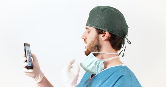Giovane medico chirurgo con barba lunga con lo smartphone si fa i video selfie con il telefonino, sfondo bianco.