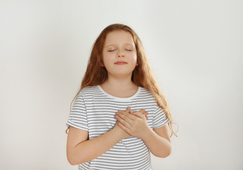 Obraz premium Cute grateful little girl with hands on chest against light background