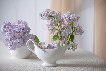 Fototapeta na wymiar Room interior with gentle purple lilacs flower blossom in jug and tea cup on table and curtain hanging on wall, tender romantic spring home decor in morning light, decorating house with syringa.