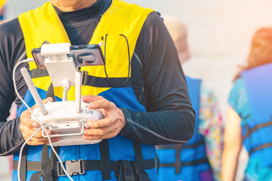 Young man in blue and yellow life vest controlling a drone to take pictures of ocean coast with tourists while traveling on a raft in the sea. Hands holding drone remote controller.