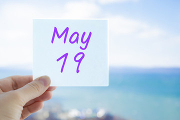 May 19th. Hand holding sticker with text May 19 on the blurred background of the sea and sky. Copy space for text. Month in calendar concept