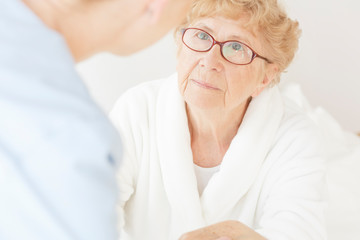 Senior lady wearing white bathrobe and glasses looking at her helpful nurse