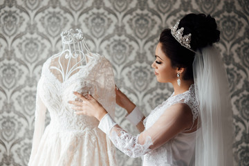 Beautiful bride with makeup and hair style. Sexy bride posing in luxury interior. Wedding morning of the bride. Bride in beautiful robe touches the dress on mannequin at home.