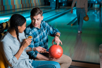 Fototapeta premium handsome boyfriend showing bowling ball to cheerful african american girl holding glass of cocktail