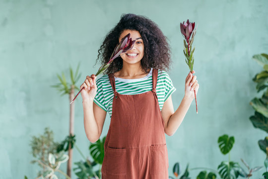 Young Beautiful Caucasian Woman In Glass Greenhouse Among Colorful Greenery Leaves And Flowers. Art Portrait Of A Girl Wearing A Shirt. Gardener, Nature Lover, Inspiration Concept.