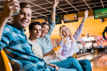 happy multicultural friends smiling at camera while sitting in bowling club with cocktails and beer