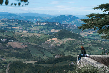 Fototapeta na wymiar Woman dressed in a motorcycle outfit. body protection turtle and knee pads. A girl is sitting on the edge of the top of the mountain. incredible view. Motorcycle travel concept Copy space. San Marino