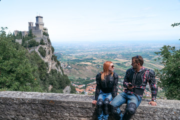 Couple of tourists have a conversation. Motorcycle outfit and sunglasses. Family vacation. The fortress, first tower on background. Summer sunny day. Top of the mountain. San Marino