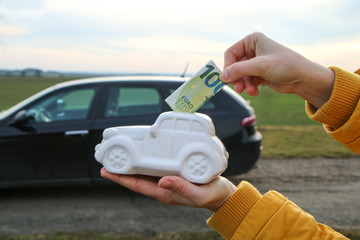 The hand is puting a banknote into the ceramic car shaped money box. The real car is behind. It can be symbol for costs for car´s repairs, investment, savings or insurance. 