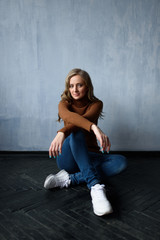 Young beautiful woman with long straight blonde hair in blue jeans and bright blouse sitting on black wooden table with crossed legs and looking in camera.