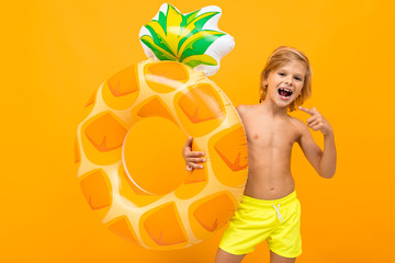 Handsome boy in swimming trunks holds a rubber ring, smiles and gesticulates isolated on orange background