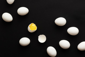 White eggs lie scattered on a black background. One egg is broken and the yolk is visible. Minimalistic design and color trend 2020. Top view