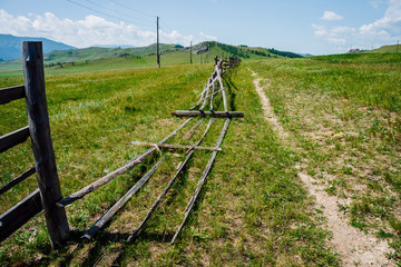 Pathway along broken wood fence and poles with wires in mountains in sunny day. Beautiful sunny alpine landscape with footpath along field behind long fence in highlands. Vivid mountain scenery. © Daniil