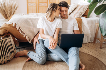 Home leisure of couple in love. Stylish attractive couple sitting on the floor near the bed in their bedroom using the Internet in a laptop