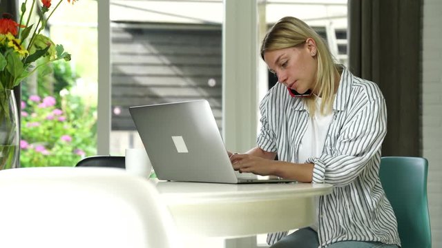 Serious caucasian female freelancer working remotely at home interior using laptop computer for making booking on web pages and support customers via mobile phone conversation