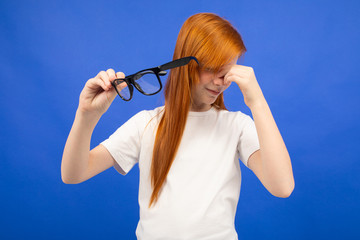 red-haired teenager girl squints while holding glasses in her hand on a blue studio background