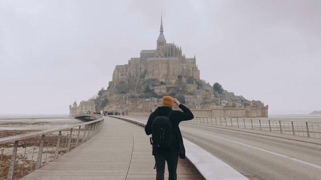 Camera follows happy excited tourist man with backpack walking to epic Mont Saint Michel castle in Normandy slow motion.