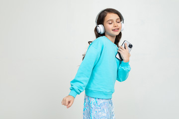 young girl in a turquoise blouse listens to music with headphones holding a phone on an isolated studio background with copy space