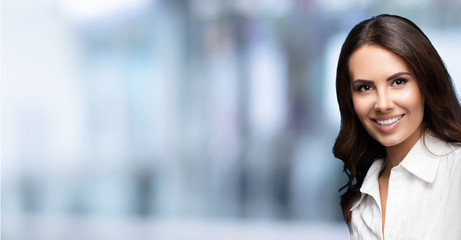 Obraz premium Portrait of happy smiling beautiful young brunette business woman, over blured office background, with blank copy space for some slogan or text. Horizontal banner composition picture.