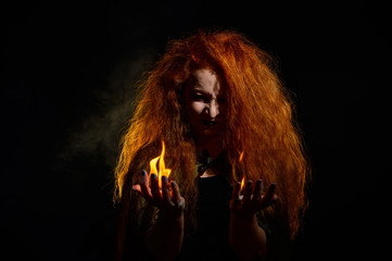 A witch with long curly hair holds a magic fire. Tongues of flame on the palms of a red-haired woman.