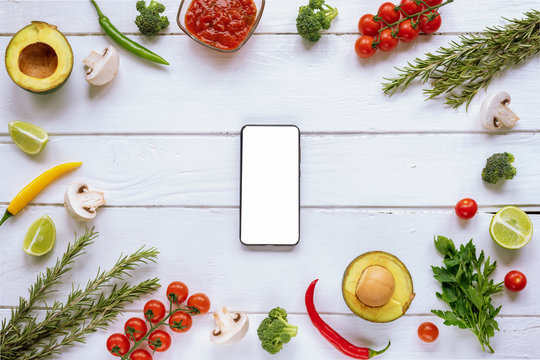 Ingredients for Italian food: tomatoes, mushrooms, broccoli, chilli peppers, avocado, rosemary and parsley on a white wooden background.  Mobile phone with blank screen Top view, flat lay, copy space.