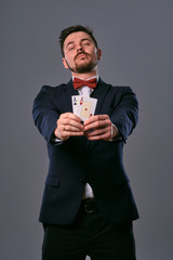 Man in black classic suit and red bow-tie showing two playing cards while posing against gray studio background. Gambling, poker, casino. Close-up.