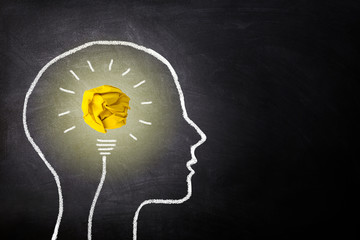 Business Idea Concept : Yellow crumpled paper ball light bulb lighting grow in brain white chalk doodle on chalkboard.