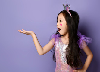 Surprised asian kid girl princess in party dress and crown is holding something on her open palm looking at it © Dmitry Lobanov