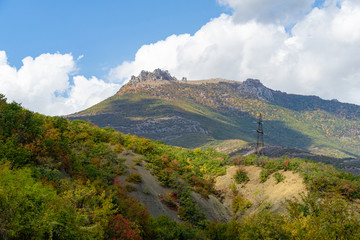 Picturesque autumn slopes of Crimean mountains on Southern coast of Crimea. Power transmission tower with wires. In distance you can see famous Demerdji mountain with clouds on blue sky.