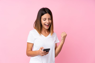 Young woman over isolated pink background with phone in victory position