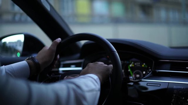 Cropped view from back seat in slow motion of business man holding hands on wheel driving modern automobile on urban city road follow rules and safety during transportation. Confidence in steering