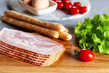 Tradition  Pancetta bacon served with pickled olives, tomatoes, green salad leaves, eggs and grissini bread, ingredients for breakfast. Close up, top view on white background