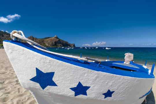 Close up of a white and blue fishing boat decorated with stars on its prow, lying ashore on a sandy beach in a sunny summer day