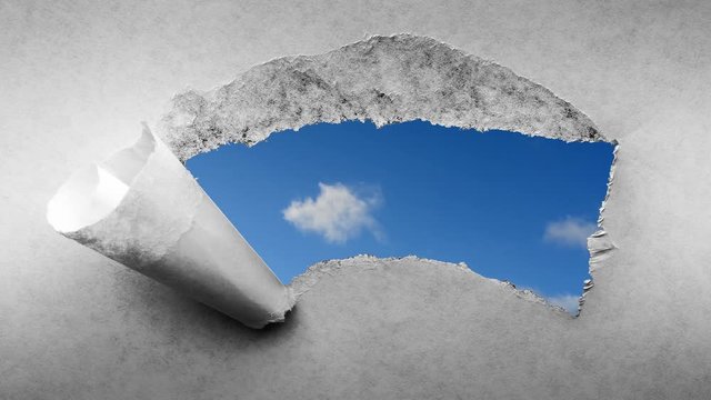 Creative 4k time laps video of fast moving clouds in the blue sky that are visible through a hole with torn edges in old retro grunge vintage paper.