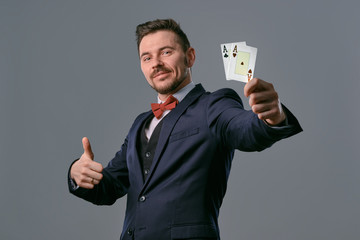 Man in black classic suit and red bow-tie showing two playing cards while posing against gray...