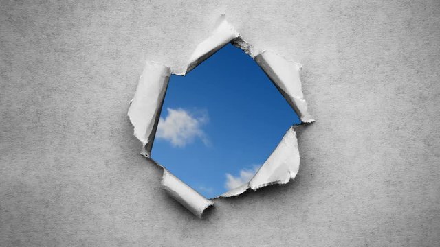 Creative 4k time laps video of fast moving clouds in the blue sky that are visible through a hole with torn edges in old retro grunge vintage paper.