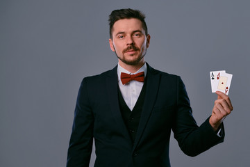 Man in black classic suit and red bow-tie showing two playing cards while posing against gray studio background. Gambling, poker, casino. Close-up.