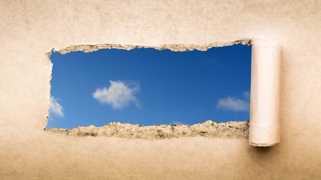 Creative 4k time laps video of fast moving clouds in the blue sky that are visible through a hole with torn edges in old retro grunge vintage paper.