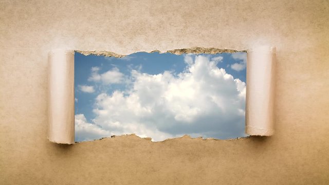Creative 4k time laps video of fast moving clouds in the blue sky that are visible through a hole with torn edges in old retro grunge vintage paper.