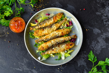 Grilled small fish in a gray plate on a black, dark concrete background. Photo of food. Fried Fish, lettuce salad, parsley, sweet and sour sauce. Top view, place for text.