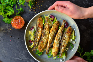 Grilled fish in a gray plate. Small fried fish and salad dressing. Top view. Wood background. Woman holds a plate of food in her hands.