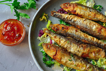 Grilled small fish in a gray plate on a gray concrete background. Food photo, top view. Fried Fish, lettuce salad, parsley, sweet and sour sauce.