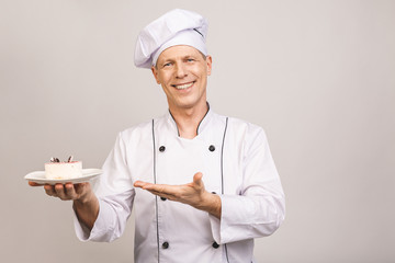 Portrait of a happy senior male chef dressed in uniform holding plate with piece of cake and looking at camera isolated over gray background.