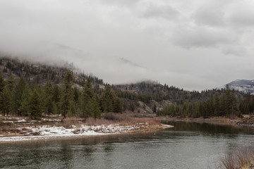 Wide and calm river in front of a tree covered mountain with snow and fog
