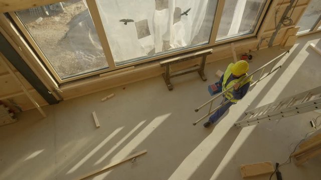 SLOW MOTION, TOP DOWN: Builder leaves the construction site with his toolbox and ladder in hand. Worker coming to work carries his metal toolbox and ladder across the room of a prefabricated house.