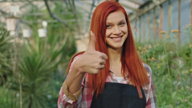 Redhead beautiful gardener in a flower greenhouse looking straight to the camera smiling large and showing a big like